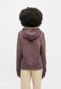 Puha anyagból készült barna kapucnis pulóver, laza szabással, kapucnival, bordázott mandzsettával és szegéllyel. A háttér egyszínű fehér.