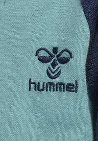 Grön stickad tyg med mörkblå detaljer, med en broderad logotyp av två vingar ovanför stiliserade blad och ordet "hummel."