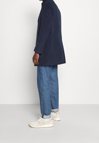 Giacca in velluto blu navy con forma sagomata, jeans blu e sneakers bianche con dettagli grigi. Bottoni visibili sulle maniche della giacca.