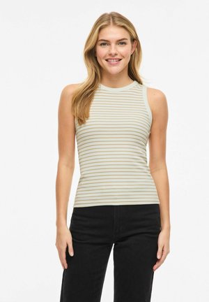 Femme souriante aux longs cheveux blonds, portant un haut sans manches beige et blanc à rayures et un pantalon noir, debout devant un fond blanc.