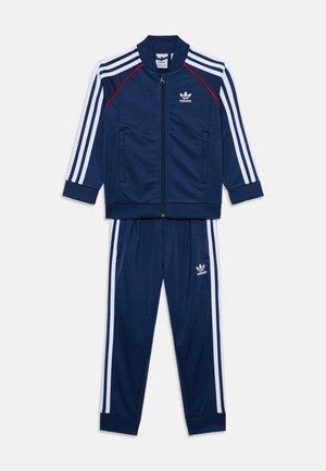 Sötétkék adidas tréningruha fehér háromcsíkos mintával. Zipzáras dzseki és rugalmas derekú nadrág jellemzi. Puha, texturált anyag.