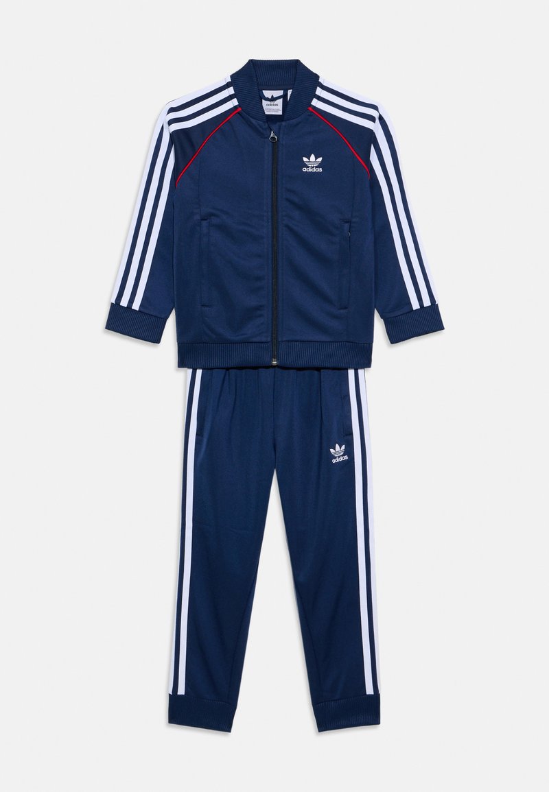 Tummansininen adidas-treenausasu, jossa on valkoinen kolmiraidan muotoilu. Mukana on vetoketjulla varustettu takki ja joustava vyötärö housuissa. Pehmeä, tekstuuri kangas.