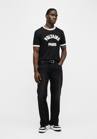 Άνδρας που φοράει μαύρο t-shirt Voltaire Paris, μαύρο τζιν, μαύρη ζώνη, λευκά και μαύρα αθλητικά παπούτσια, και ένα κολιέ με πέρλες, στέκεται μπροστά σε απλό φόντο.