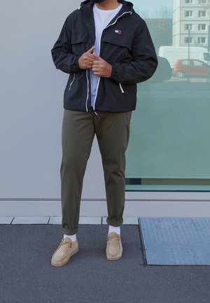 Personne portant une veste noire zippée, un t-shirt blanc, un pantalon vert olive, des chaussettes blanches et des chaussures en daim beige, debout sur le trottoir devant un bâtiment.