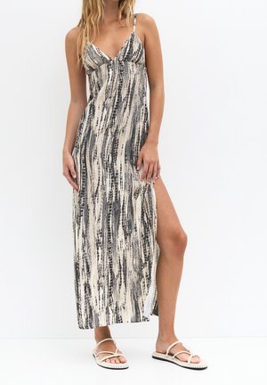 Kvinde iført ærmeløs maxikjole med sorte og beige abstrakte lodrette striber og højt sidespalt, kombineret med hvide sandaler.
