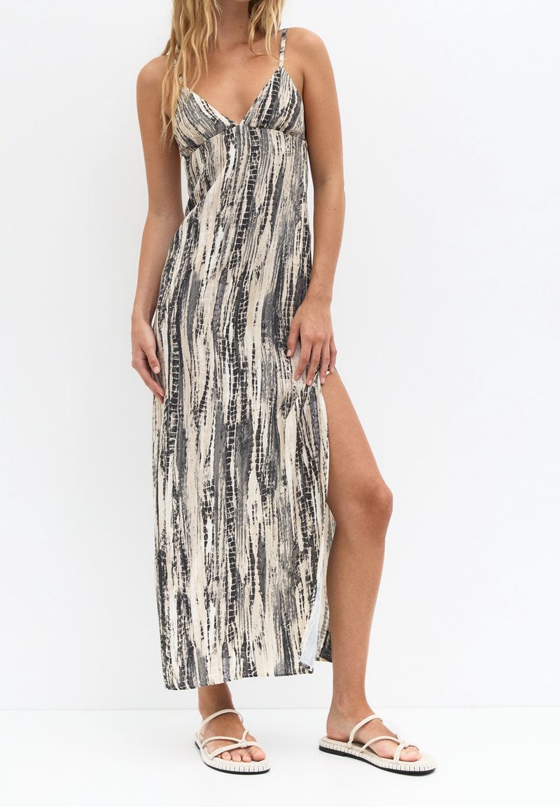 Femme portant une robe maxi sans manches avec des rayures verticales abstraites noires et beiges, fendue sur le côté jusqu'à la cuisse, associée à des sandales blanches.