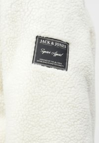 Veste en polaire blanche avec une surface texturée et une étiquette noire. L'étiquette indique "JACK & JONES", avec un logo et des détails de conception.