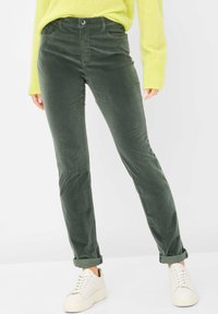 Grüne Cordhosen mit schlanker Passform, ausgestattet mit Fronttaschen und aufgerollten Säumen, kombiniert mit weißen Sneakern und einem leuchtend gelben Pullover.