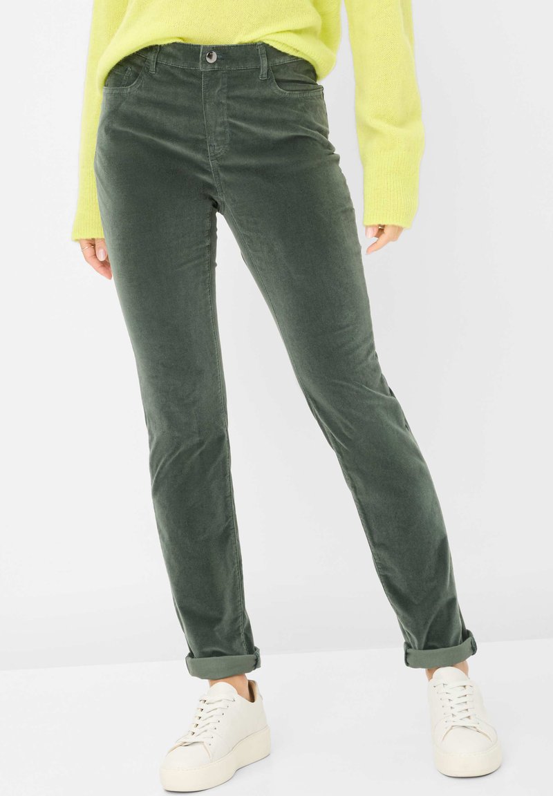 Grüne Cordhosen mit schlanker Passform, ausgestattet mit Fronttaschen und aufgerollten Säumen, kombiniert mit weißen Sneakern und einem leuchtend gelben Pullover.