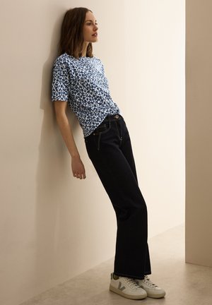 T-shirt a fantasia floreale blu e bianca, jeans in denim neri e sneakers bianche con dettagli verdi. Il modello si appoggia a un muro.
