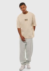 Beige oversized t-shirt met "DROPSIZE" in bruin gedrukt, gecombineerd met lichtgroene baggy jeans en witte sneakers. Soepele, casual stof.