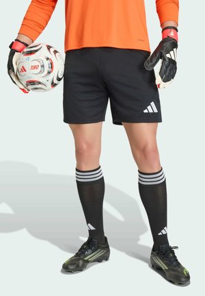 Bramkarz piłkarski ubrany w pomarańczową koszulkę, czarne spodenki, rękawice i korki, trzymający biało-czarną piłkę do piłki nożnej Adidas.