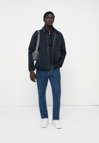 Navy jacket with a snap front, layered over a dark top, paired with blue jeans and white sneakers. Small grey crossbody bag on shoulder.
