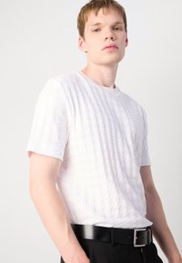 Cămașă albă cu mânecă scurtă, cu un model texturat în grilaj și guler rotund, asociată cu o curea neagră și pantaloni.