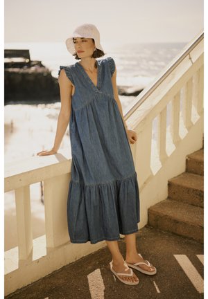 Denim maksi kleita ar V-veida kakla izgriezumu, krokotām plecu daļām un pakāpenisku apakšmalu. Valkāta ar baltām flip-flop sandalēm un tamborētu rozā cepuri. Pludmales fons.