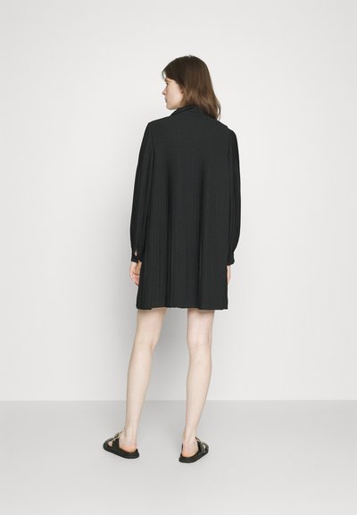 Rochie neagră, plisată, cu mâneci bufante lungi și guler înalt. Design simplu, se termină deasupra genunchilor, asortată cu sandale plate.