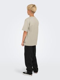 Beige t-shirt med korte ærmer og en afslappet pasform, parret med løse sorte bukser og sorte sneaks med hvide detaljer, set fra ryggen.