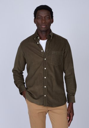 Chemise en velours côtelé vert olive foncé avec un col boutonné, une poche poitrine simple et des boutons blancs ; portée par-dessus un t-shirt blanc.