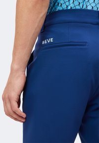 Personne portant un pantalon bleu avec le mot "SEVE" au-dessus d'une poche arrière, debout avec la main gauche visible et une partie d'une chemise bleue à motifs.