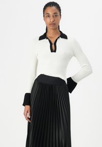 White ribbed top with black accents, featuring a collar and a zip detail. Paired with a black pleated skirt with a logo waistband.