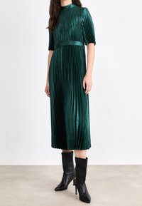Groene geplooide midi-jurk met korte mouwen en een tailleband. Gemaakt van zachte stof, gestyled met een hoge hals en gecombineerd met zwarte haklaarzen.