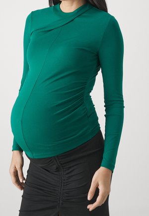 Personne enceinte portant un haut vert ajusté à manches longues et une jupe noire froncée, debout devant un fond clair uni.