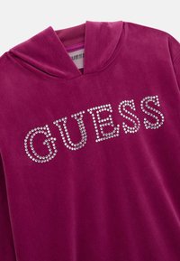 Fuksijos aksominis džemperis su reguliuojamu kapišonu ir deimantų "GUESS" logotipu priekyje. Minkšta tekstūra ir laisvo kirpimo dizainas.