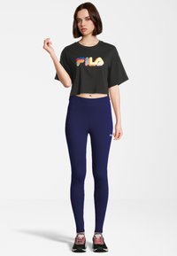 Camiseta corta negra con el logo "FILA" multicolor, combinada con leggings azul marino. La modelo lleva zapatillas negras con detalles rosas.