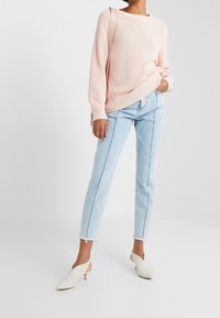Pull en tricot rose clair à manches longues avec boutons sur le côté, associé à un jean taille haute bleu clair et des mules couleur crème.
