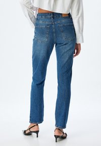 Siniset farkut suoran lahkeen leikkauksella, joissa on kaksi takataskua ja nahkapattilenkkiriipus vyötäröllä. Rakoileva kangas, jossa saumat näkyvät.