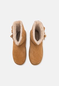 Paire de bottines en daim beige avec doublure en fourrure crème et nœuds latéraux, vues de dessus sur fond blanc.