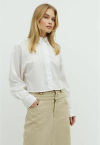 Camicia bianca a righe con maniche lunghe, abbinata a una gonna beige. La camicia ha un colletto classico e una vestibilità comoda.