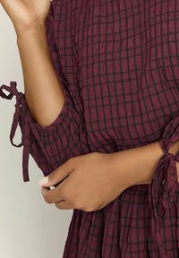 Arms crossed over torso wearing a crinkled dark purple checkered dress with tied sleeve cuffs.