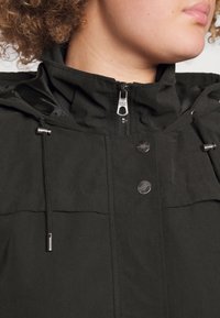 Giacca nera con cappuccio, dotata di cordini regolabili, bottoni a scatto e chiusura con zip. Presenta una texture liscia e un design minimalista.
