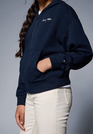 Osoba z warkoczami, ubrana w granatową bluzę Tommy Hilfiger na zamek oraz jasne spodnie, z ręką w kieszeni bluzy.