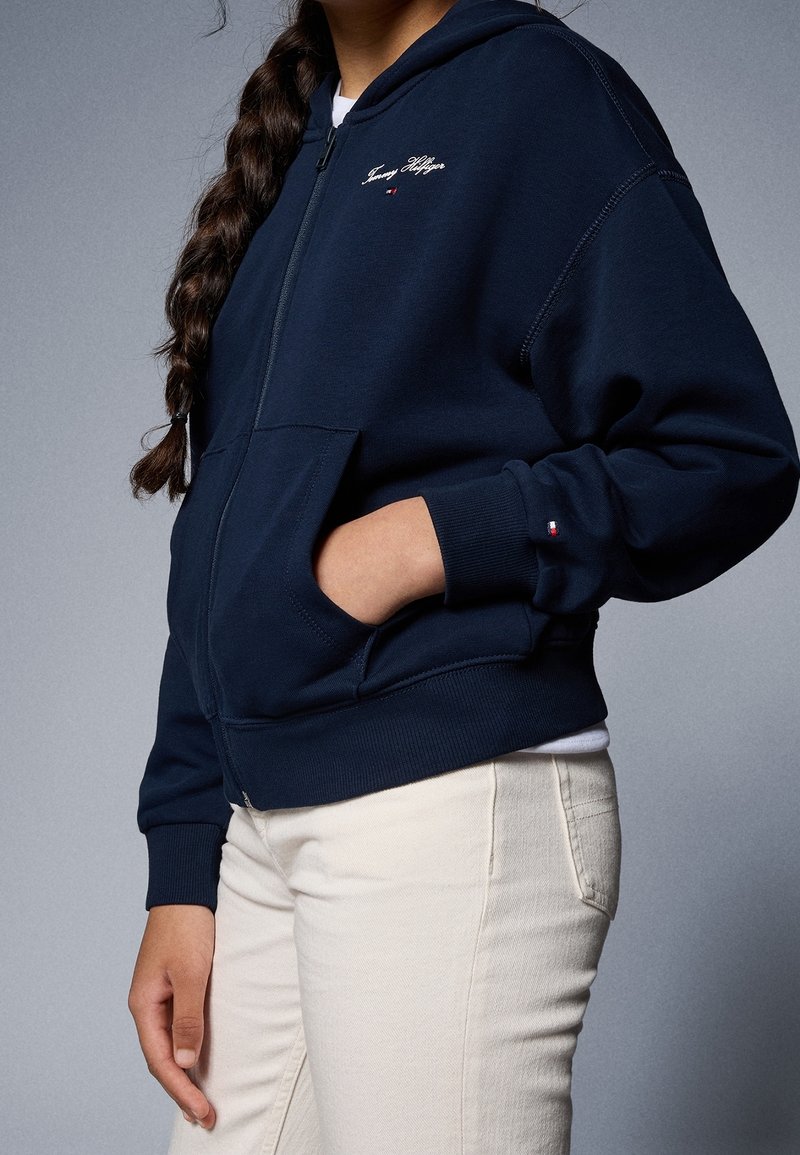 Personne aux cheveux tressés portant un sweat à capuche zippé bleu marine Tommy Hilfiger et un pantalon de couleur claire, la main dans la poche du sweat.