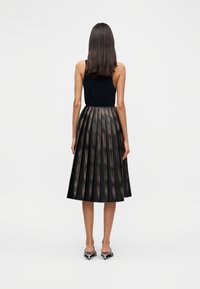 Γυναίκα που στέκεται γυρισμένη πλάτη, φορώντας μαύρο αμάνικο τοπ, μεταλλική πλισέ μέση φούστα και μαύρα σανδάλια με χαμηλό τακούνι σε λευκό φόντο.