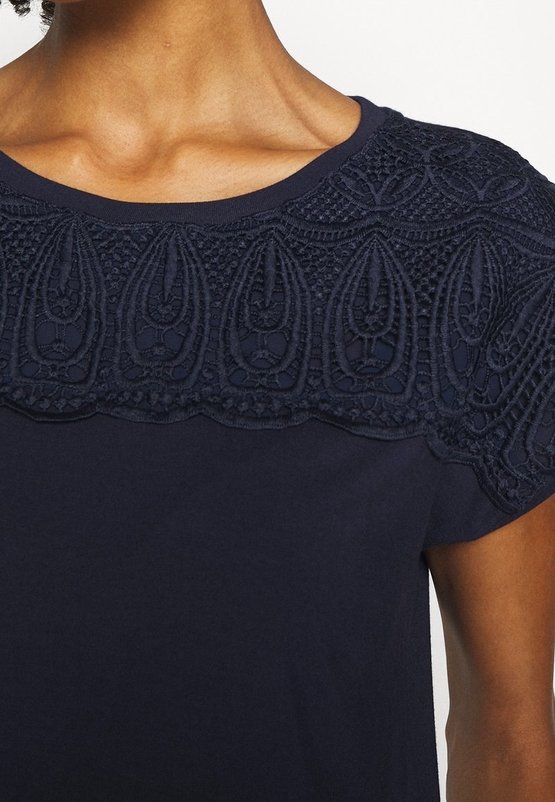 Detailní záběr osoby v námořnické modré blůze s propracovanou krajkovou výšivkou podél výstřihu a ramen.