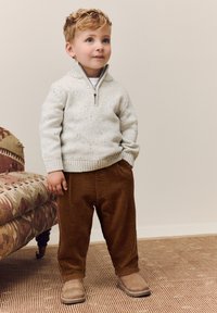 Svetlosivý pletený sveter s golierom na zips, posiaty bodkami; hnedé manšestrové nohavice; hnedé semišové čižmy. Stojí na vzorovom koberci vedľa látkovej pohovky.