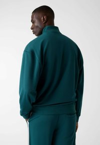 Groene, gestructureerde jas met rits en een hoge kraag, voorzien van lange mouwen en een ontspannen pasvorm, gezien vanaf de achterkant met bijpassende broek.