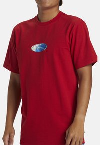 Camiseta de algodón roja con mangas cortas que presenta un logo circular en azul y blanco en el pecho. Diseño clásico de cuello redondo.