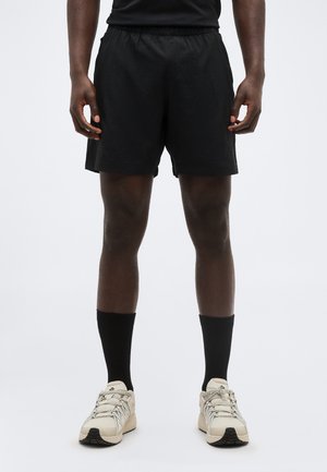 Homme portant un short de sport noir, des chaussettes noires classiques et des chaussures de course beiges, debout avec les bras détendus le long du corps sur un fond blanc.