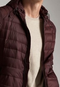 Bordeaux quiltet pufferjakke med hætte, med lynlåslukning og snoretræk, båret over en lys beige skjorte.