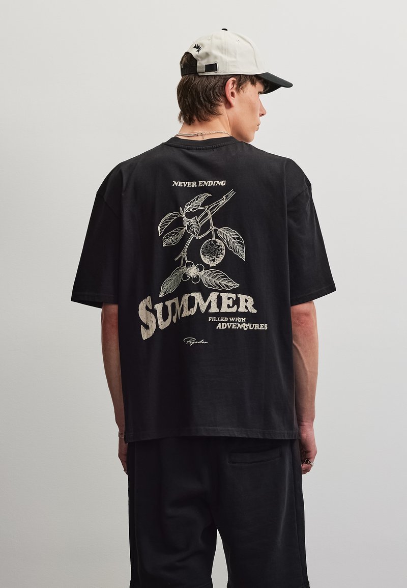 Νέος άνδρας που φοράει μαύρο T-shirt και λευκό καπέλο μπέιζμπολ, στην πλάτη του T-shirt υπάρχει εκτύπωση με βοτανικό σχέδιο και κείμενο «Never Ending Summer γεμάτο περιπέτειες.»