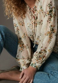 Personne aux cheveux bouclés portant un blouse florale transparente avec un col à nouer, un jean bleu et des chaussures plates dorées, assise les jambes croisées.