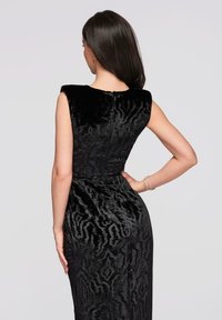 Femme aux longs cheveux noirs portant une robe en velours noire à motifs sans manches, main sur la taille, dos tourné sur un fond gris clair.