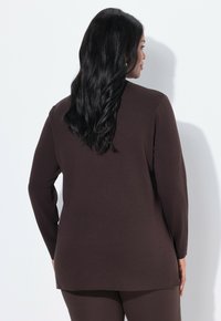 Femme aux longs cheveux noirs portant un haut à manches longues marron foncé et un pantalon assorti, debout contre un mur blanc, de profil.