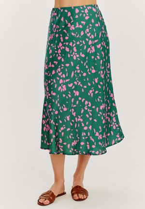 Zielona spódnica midi z rozproszonymi różowymi abstrakcyjnymi plamami, noszona z brązowymi płaskimi sandałami, dolne partie nóg widoczne na jednolitym tle.