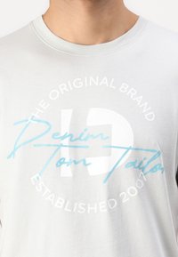 Lysegrå bomullst-skjorte med rund hals. Frontdesignet inkluderer hvit og lyseblå tekst med "Denim Tom Tailor" og "Etablert 2007."