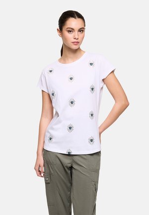 Joven con el pelo recogido lleva una camiseta blanca con patrones de ojos azules y pantalones cargo verde oliva, de pie frente a un fondo liso.